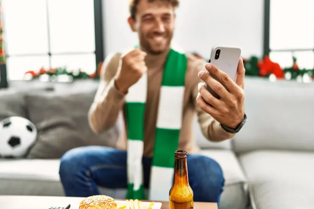 Torcedor sentado no sofá comemora enquanto olha o celular, usando cachecol do time, com lanche e cerveja à frente.