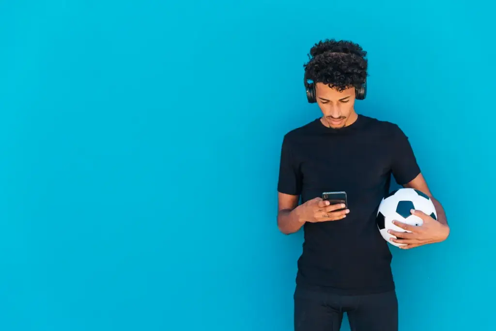Homem jovem segurando bola de futebol enquanto usa o celular com fones de ouvido, em fundo azul.