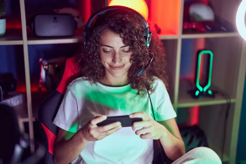 Mulher sorrindo enquanto joga no smartphone usando headset gamer em ambiente iluminado por luzes coloridas.