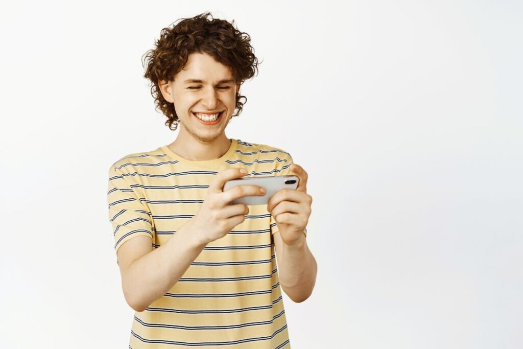 Jovem sorrindo usando o celular com as duas mãos em fundo branco neutro