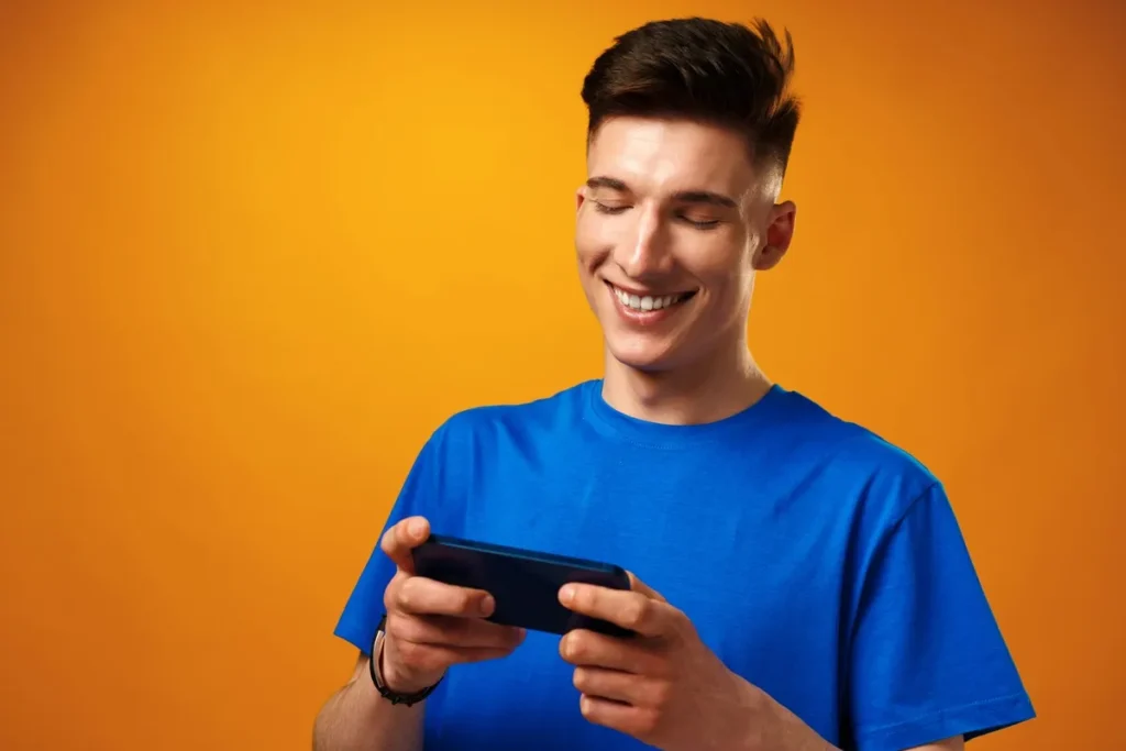 Jovem usando camiseta azul sorrindo enquanto joga no celular, segurando o smartphone com as duas mãos diante de um fundo laranja.
