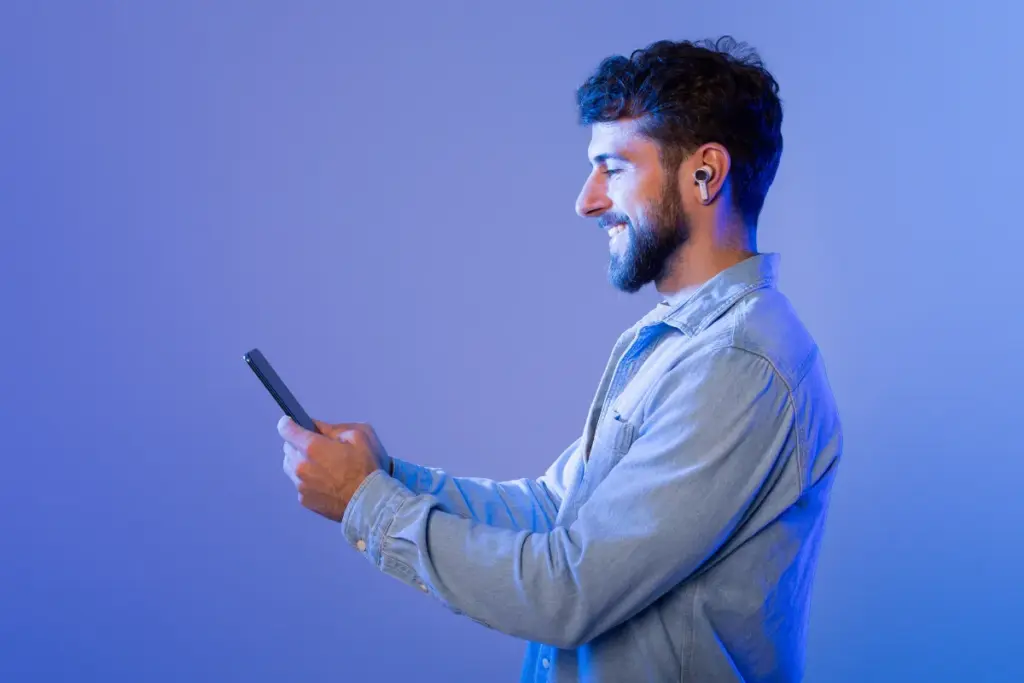 Homem sorrindo enquanto segura um smartphone e usa fones de ouvido sem fio em um ambiente iluminado em azul.