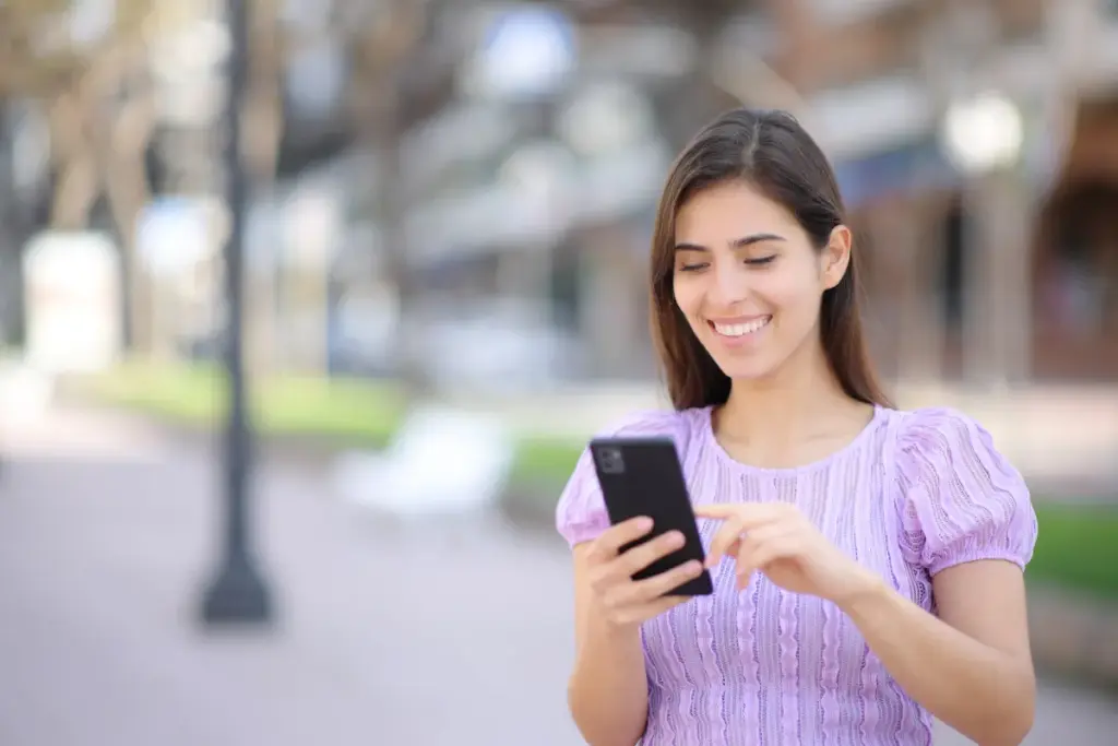 Mulher sorrindo enquanto segura um smartphone e toca na tela em um ambiente externo desfocado.