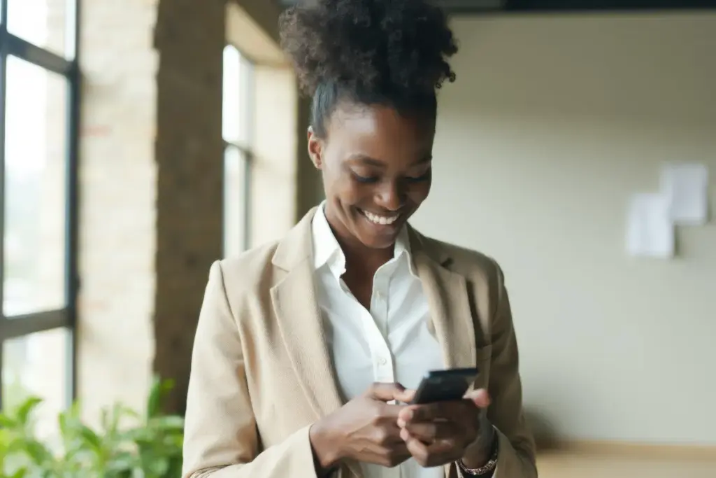 Mulher sorrindo enquanto segura um smartphone com as duas mãos em um ambiente interno iluminado.