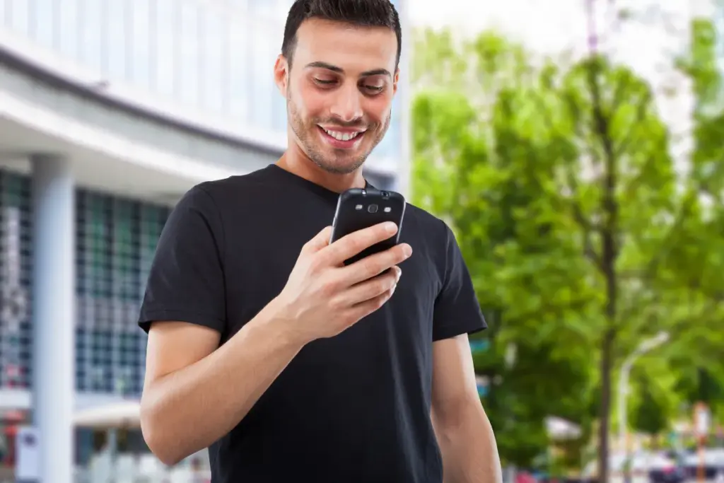 Homem sorrindo enquanto segura um smartphone e olha para a tela em um ambiente externo com árvores ao fundo.