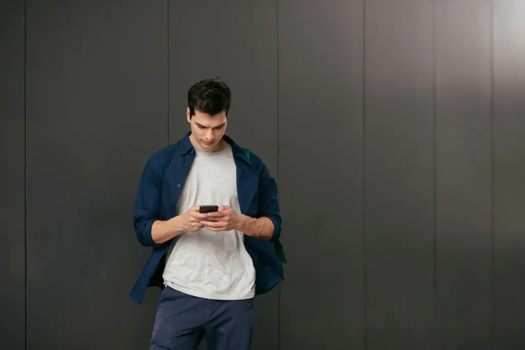 Homem caminhando enquanto olha para a tela do smartphone que segura com as duas mãos diante de um fundo escuro.