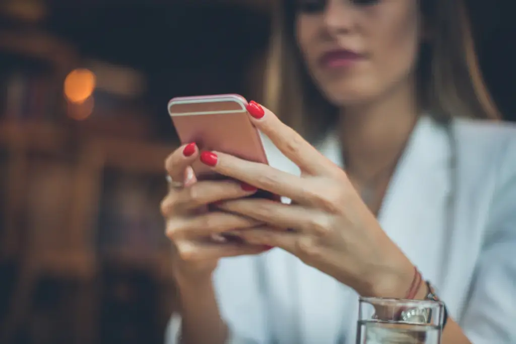 Mulher segurando um smartphone enquanto digita na tela, com unhas pintadas de vermelho.