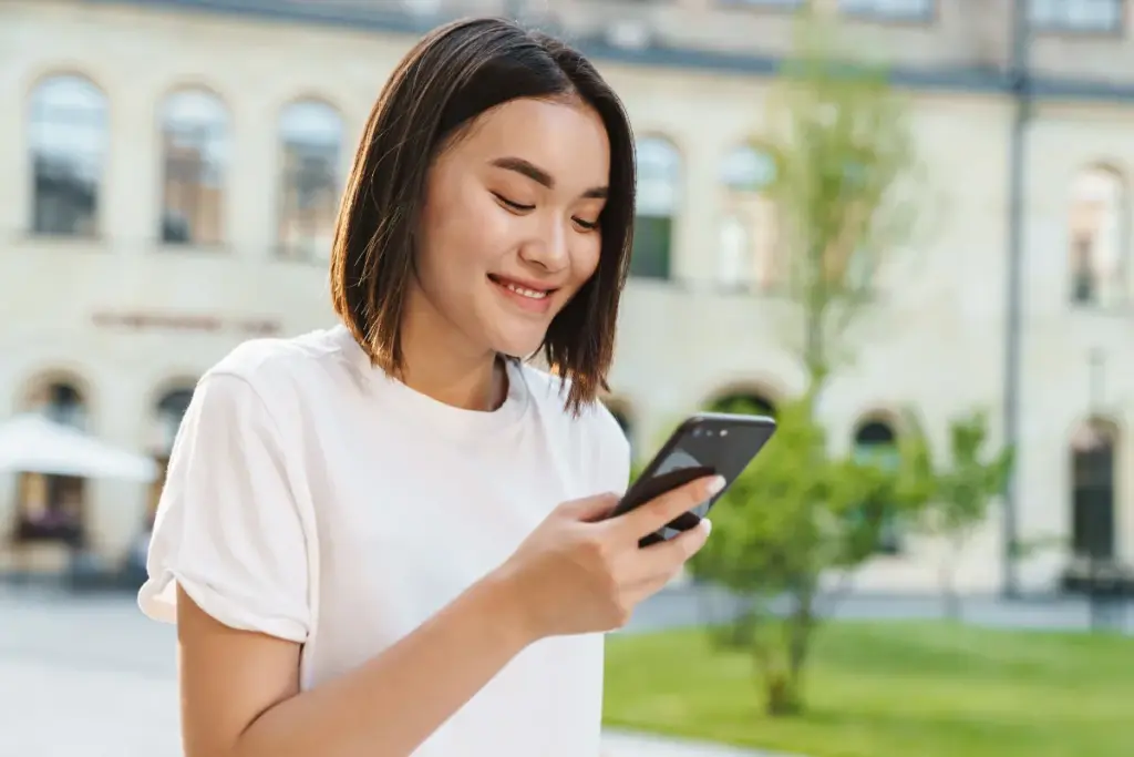 Mulher sorrindo enquanto segura um smartphone e observa a tela em um ambiente externo.