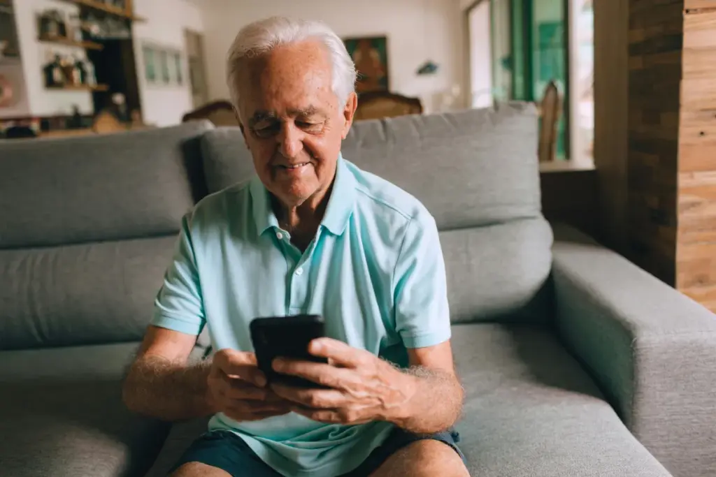 Homem idoso sentado no sofá segurando um smartphone com as duas mãos enquanto olha para a tela.