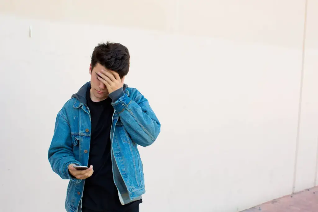 Jovem usando jaqueta jeans olhando para o celular enquanto leva a mão à testa com expressão de preocupação.