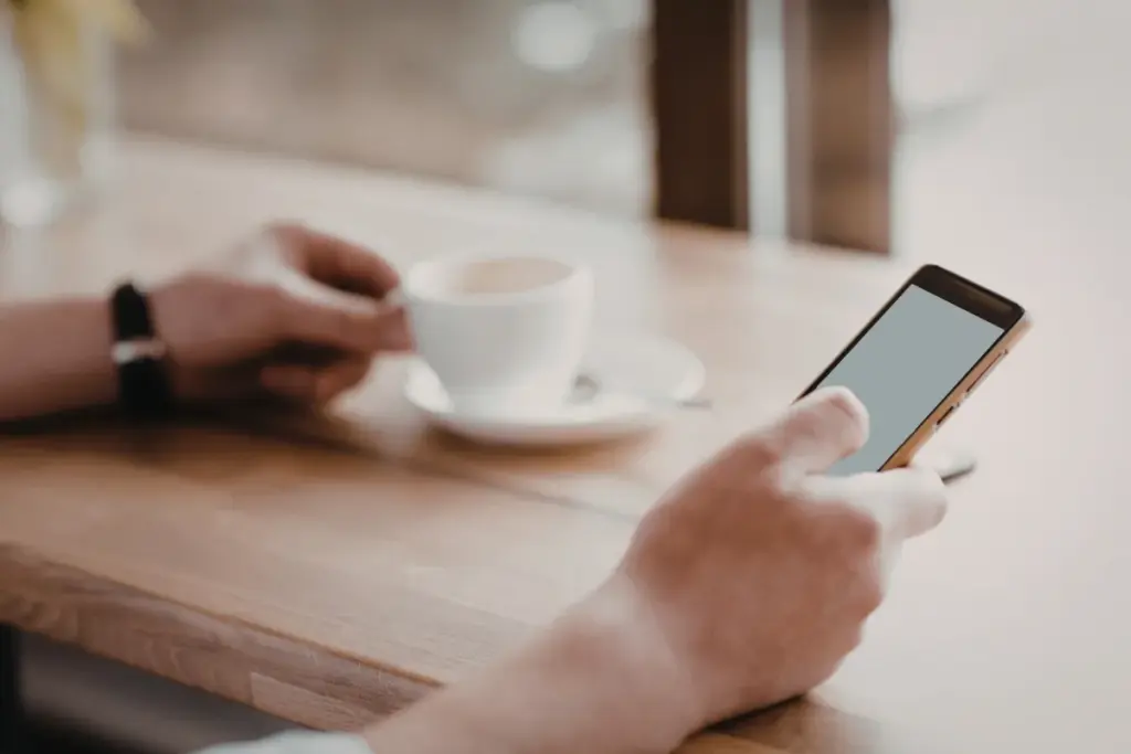 Pessoa segurando um smartphone enquanto está sentada à mesa com uma xícara de café ao lado.