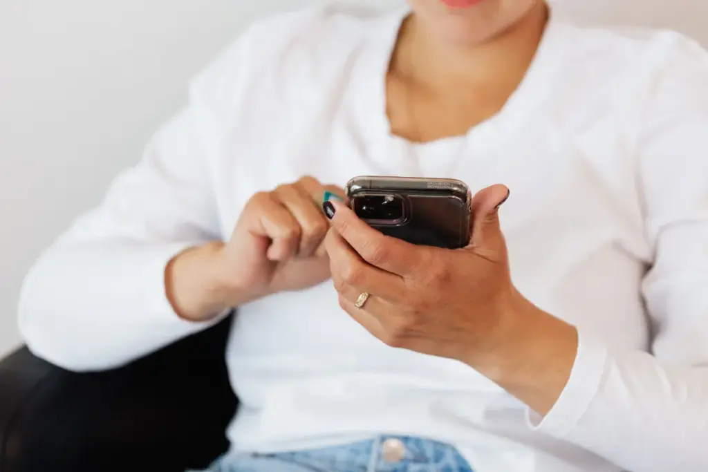 Pessoa vestindo camiseta branca segurando um smartphone enquanto toca na tela com o dedo, concentrada no aparelho.