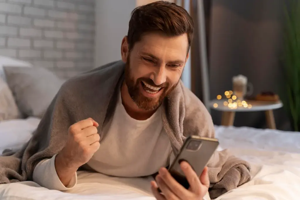 Homem sorrindo e comemorando ao usar aplicativo no celular, deitado na cama em ambiente doméstico.