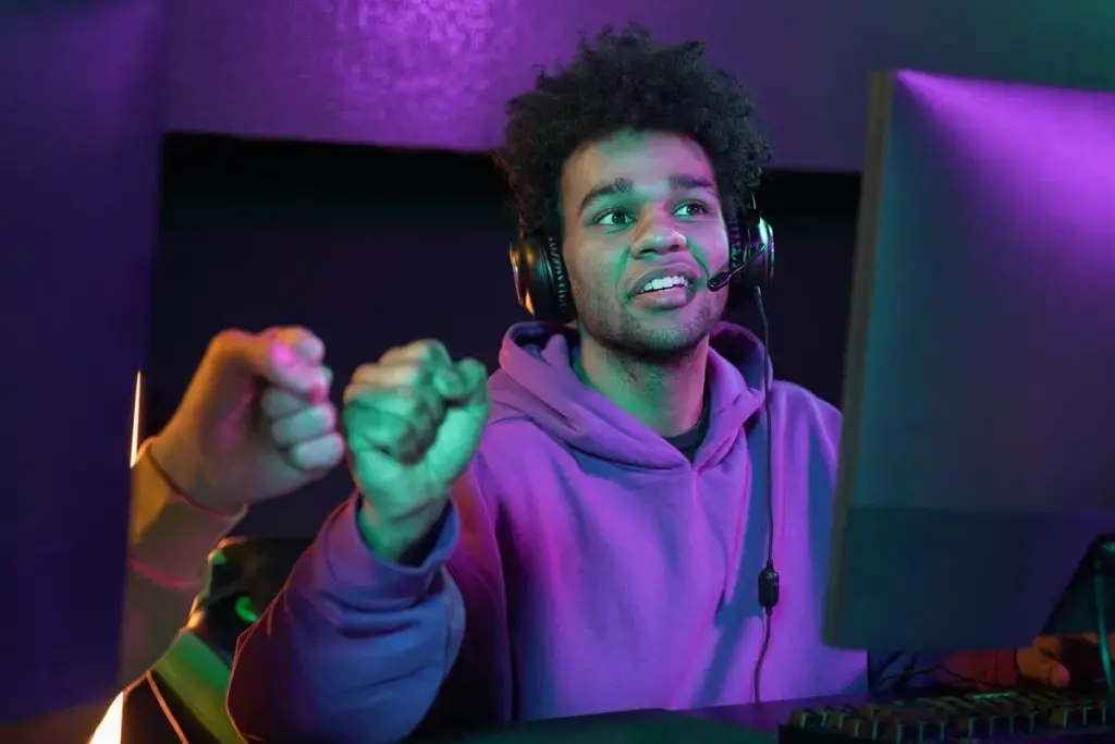 Jogador usando headset gamer comemorando vitória em jogo no computador com gesto de celebração.