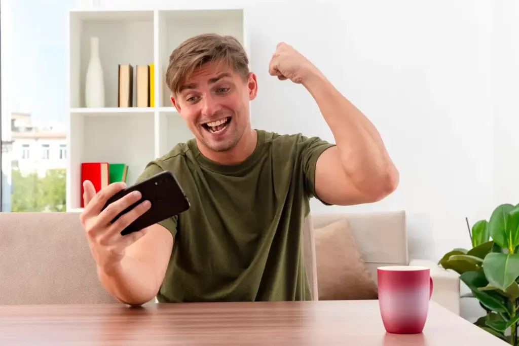 Homem sorrindo e comemorando enquanto olha para o celular em ambiente doméstico