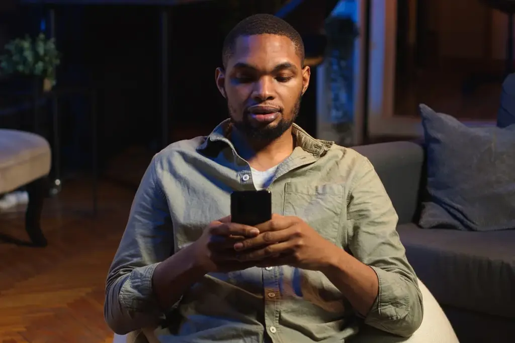 Homem sentado no sofá usando smartphone à noite, concentrado na tela, em ambiente interno com pouca luz.