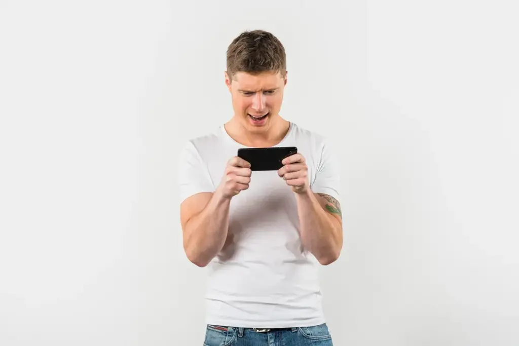 Homem jovem segurando smartphone na horizontal, demonstrando frustração enquanto joga, vestindo camiseta branca, em fundo branco