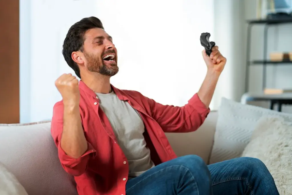 Homem sentado no sofá comemorando vitória em jogo, segurando controle de videogame e levantando o punho.