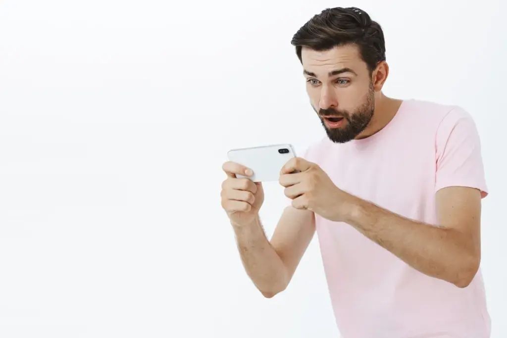Homem jovem segurando smartphone na horizontal, com expressão de surpresa enquanto joga, vestindo camiseta rosa, em fundo branco