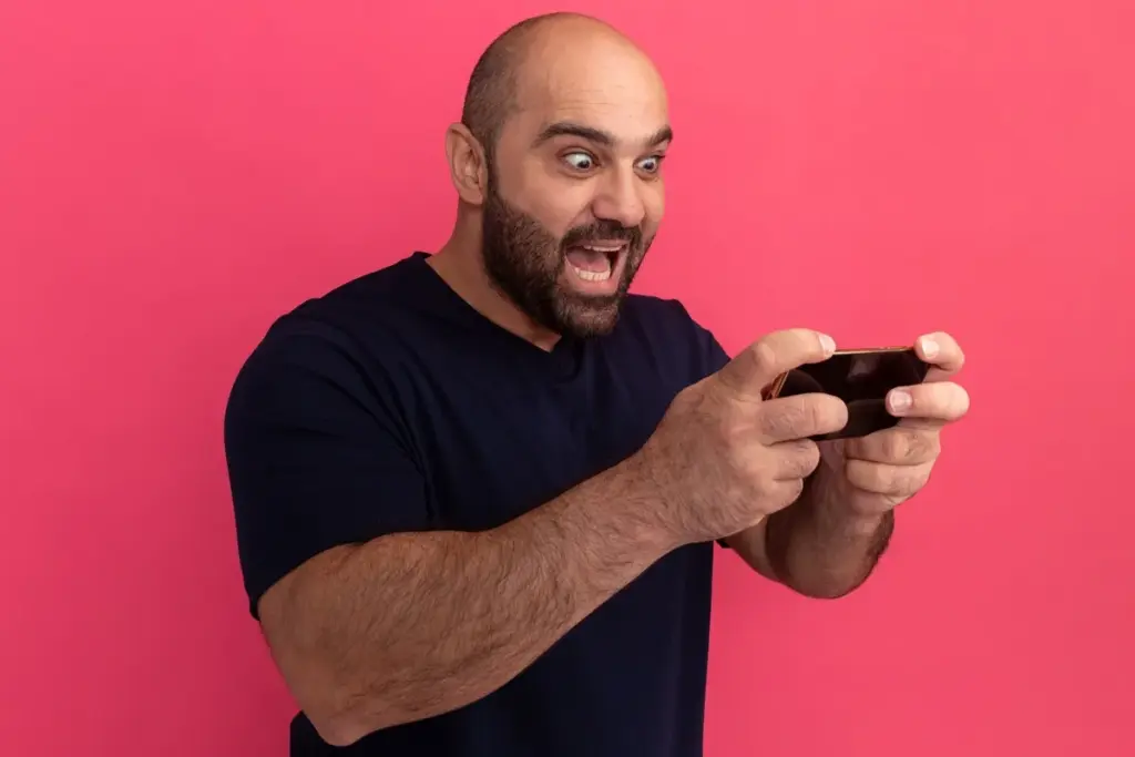 Homem careca com barba jogando no celular e reagindo com surpresa, em fundo rosa vibrante.