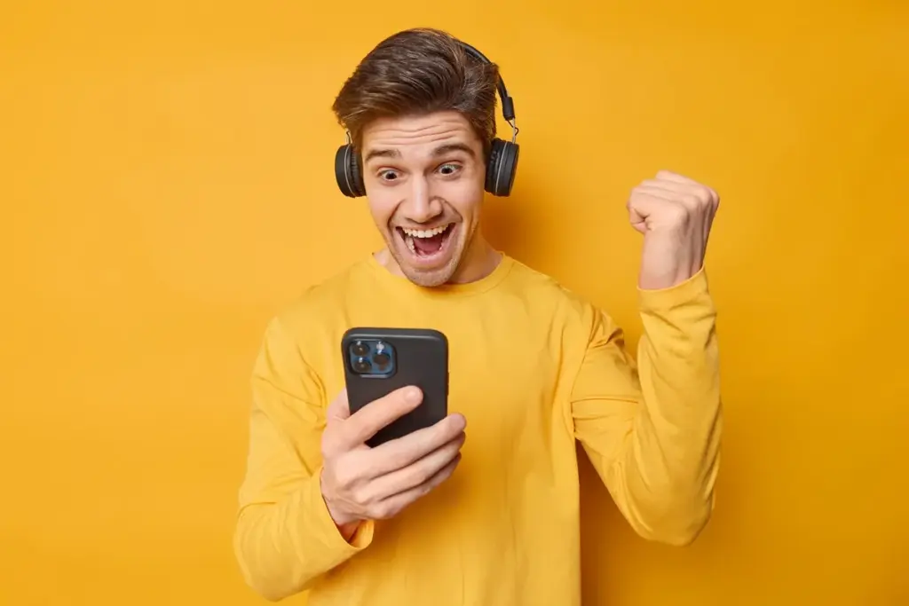 Homem sorrindo e comemorando sucesso no smartphone com fones de ouvido e punho cerrado