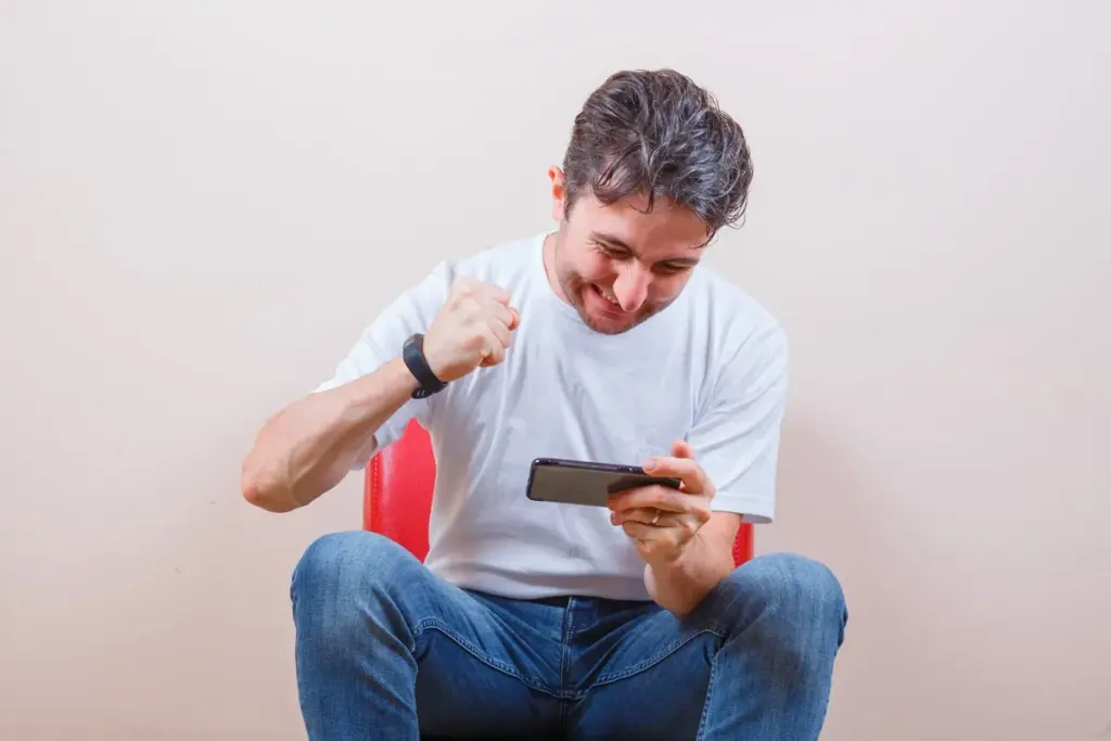 Homem sentado comemorando enquanto usa celular após comprar moedas em jogo online.