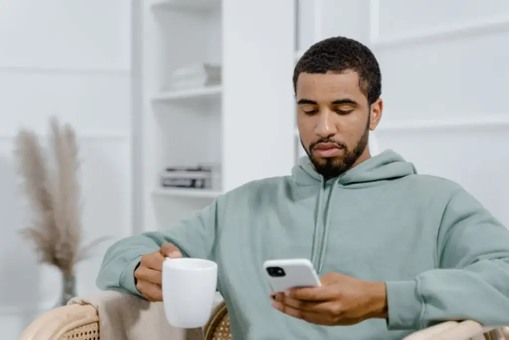 Homem sentado no sofá segurando uma xícara e usando um smartphone enquanto olha para a tela.