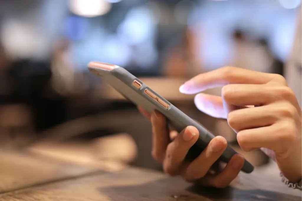 Mão segurando smartphone enquanto o dedo toca a tela iluminada em ambiente interno desfocado.