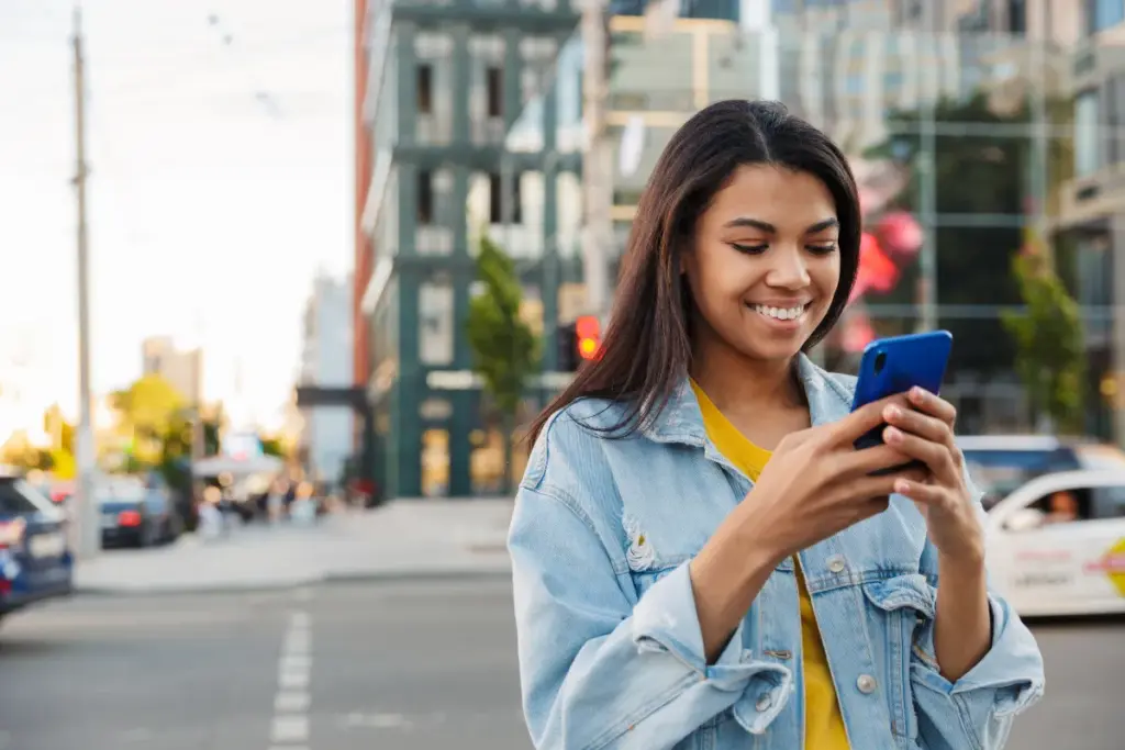 Mulher sorrindo enquanto usa o celular em uma rua da cidade durante o dia.