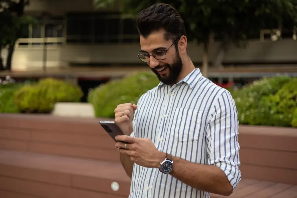 homem sorrindo e levantando o punho enquanto olha para a tela do smartphone em um espaço ao ar livre