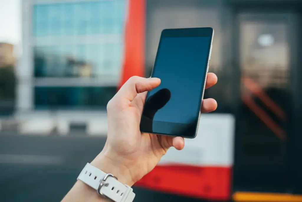 Mão segurando um smartphone com a tela escura em ambiente externo, com um prédio e elementos urbanos desfocados ao fundo.