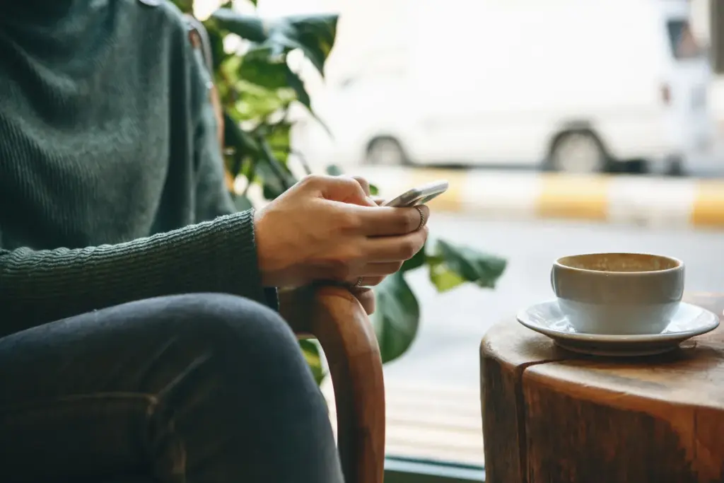 Pessoa sentada segurando um celular enquanto está em uma cafeteria, com uma xícara de café sobre a mesa ao lado.