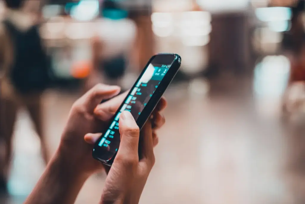 Mãos segurando um smartphone e navegando em uma interface com dados e listas na tela em ambiente interno desfocado.