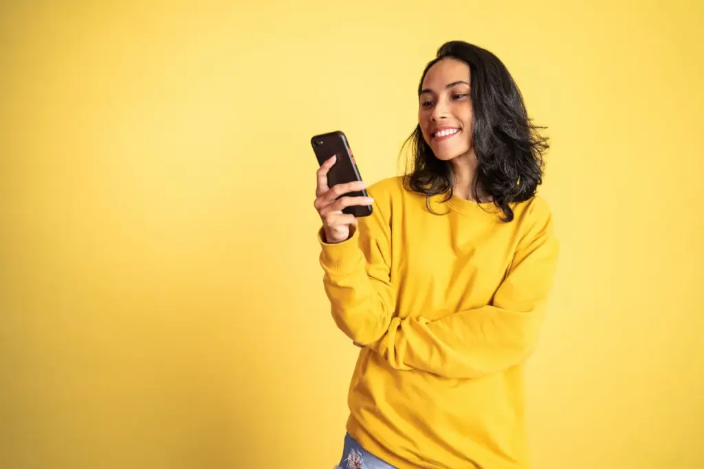 Mulher vestindo blusa amarela sorri enquanto olha para o celular em fundo amarelo.