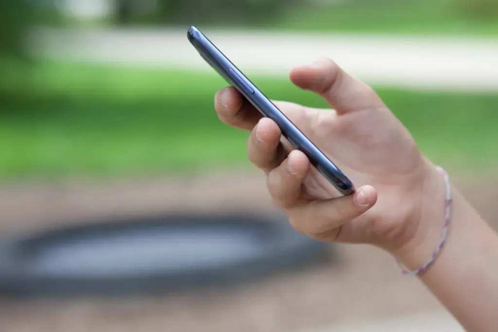 Mão segurando um smartphone em ambiente externo com área verde desfocada ao fundo.